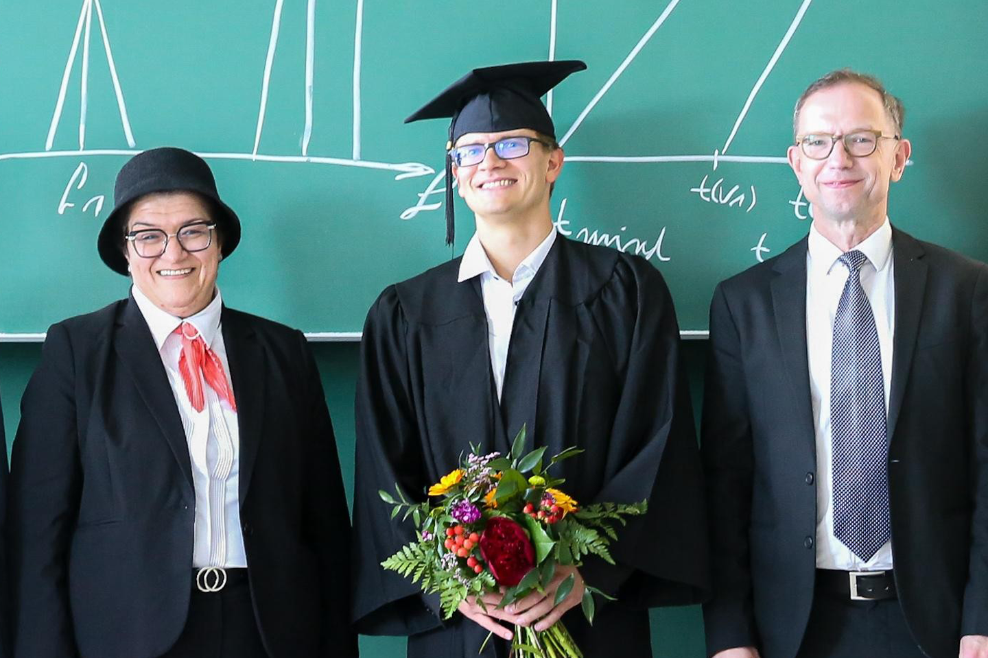 Gruppenfoto Promovend Maik Wolf in der Mitte mit Talar und Motarboard sowie Blumenstrauß