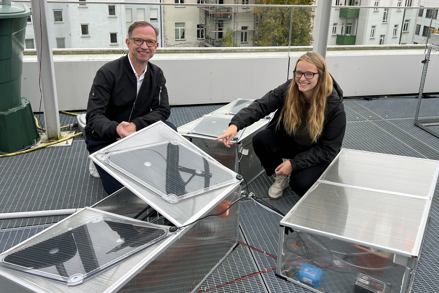 Prof. Rudolph und Laura hocken vor ihrem Versuchsaufbau mit Mini-Photovoltaikpanels auf dem Dach der Fakultät und blicken in die Kamera/Verlinkung führt zur Projektwebsite