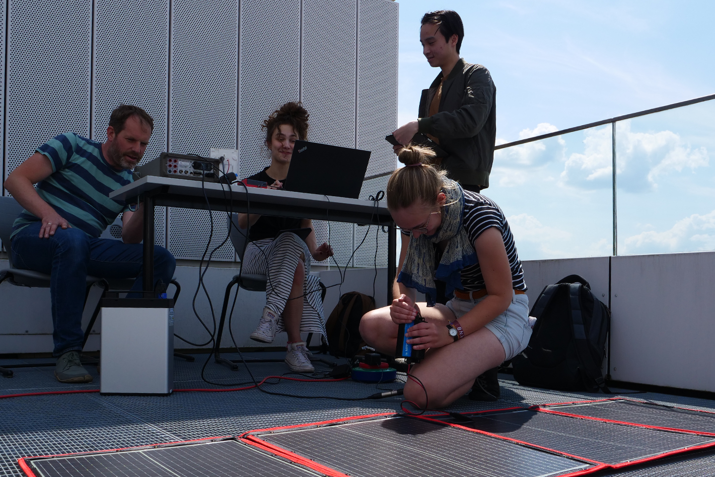 Studierende sitzen am Tisch und vor einer PhotovoltaikFolie und führen Messungen durch/Link führt zur Projektwebsite